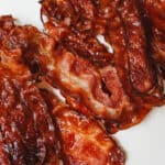 Crispy, cooked strips of bacon arranged on a white surface showcase how to cook bacon in the oven for a rich brown color and glossy, caramelized texture with slightly curled edges.
