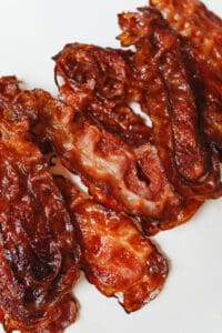Crispy, cooked strips of bacon arranged on a white surface showcase how to cook bacon in the oven for a rich brown color and glossy, caramelized texture with slightly curled edges.
