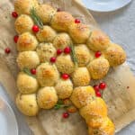 Bread rolls arranged in the shape of a Christmas tree, inspired by a Puff Pastry Christmas Tree, topped with sesame seeds, herbs, rosemary sprigs, and cranberries, displayed on parchment paper with empty white plates nearby.