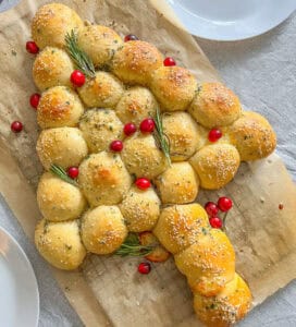 Bread rolls arranged in the shape of a Christmas tree, inspired by a Puff Pastry Christmas Tree, topped with sesame seeds, herbs, rosemary sprigs, and cranberries, displayed on parchment paper with empty white plates nearby.