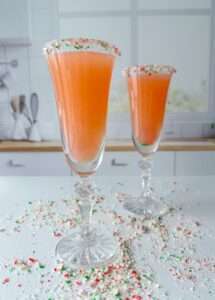 Two tall champagne flutes filled with an orange-pink Holiday Candy Cane Prosecco, each rimmed with crushed red, green, and white candy, stand on a white countertop with scattered candy. Cozy kitchen background.