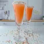 Two tall champagne flutes filled with a Holiday Candy Cane Prosecco, each glass rimmed with crushed candy pieces, stand on a white countertop in a modern kitchen. Crushed candy is scattered around the glasses.
