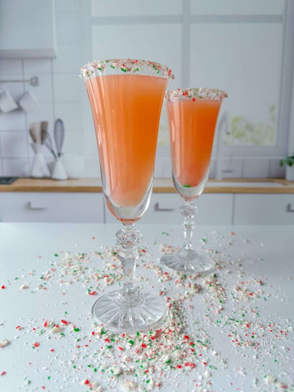Two tall champagne flutes filled with a Holiday Candy Cane Prosecco, each glass rimmed with crushed candy pieces, stand on a white countertop in a modern kitchen. Crushed candy is scattered around the glasses.