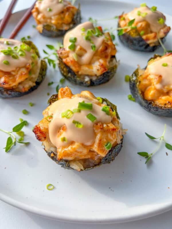 Five Chicken Bake Cups wrapped in seaweed, topped with creamy sauce and chopped green onions, are arranged on a white plate with sprigs of fresh herbs.