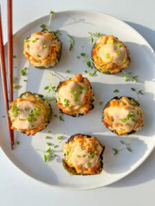 Six Chicken Bake Cups topped with creamy sauce and chopped chives are arranged on a round white plate with a few sprigs of microgreens. A pair of wooden chopsticks rests on the plate's edge.