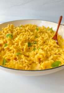 A bowl of Creamy Butternut Squash Pasta with ruffled edges, garnished with fresh green herbs, and a wooden spoon resting inside. The pasta is coated in a smooth yellow sauce.