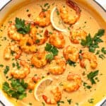 A pan filled with creamy orange sauce topped with cooked shrimp, garnished with fresh parsley and lemon slices. The shrimp are arranged attractively on the sauce, creating a visually appealing dish. Lemon wedges are placed on the side.