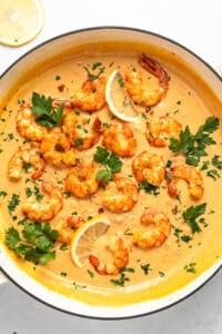 A pan filled with creamy orange sauce topped with cooked shrimp, garnished with fresh parsley and lemon slices. The shrimp are arranged attractively on the sauce, creating a visually appealing dish. Lemon wedges are placed on the side.