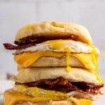 A stack of breakfast sandwiches featuring crispy bacon, melted cheese, and a fried egg between golden English muffins. The runny yolk drips down the sides. The background is softly blurred.
