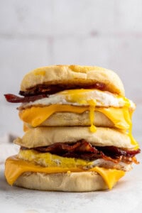 A stack of breakfast sandwiches featuring crispy bacon, melted cheese, and a fried egg between golden English muffins. The runny yolk drips down the sides. The background is softly blurred.
