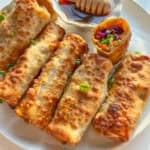 A plate of crispy Vegetable Egg Rolls garnished with chopped green onions, with one cut open to reveal a colorful veggie filling. A small dish of honey with a honey dipper is on the side.
