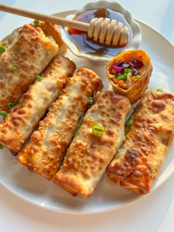 A plate of crispy Vegetable Egg Rolls garnished with chopped green onions, with one cut open to reveal a colorful veggie filling. A small dish of honey with a honey dipper is on the side.
