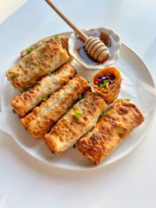 A plate of six golden-brown Vegetable Egg Rolls garnished with chopped green onions, one roll cut open to reveal colorful filling, served with a small bowl of dipping sauce and a honey dipper.