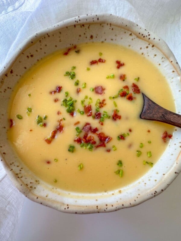 A bowl of creamy yellow soup, reminiscent of Creamy Vegetable Soup, garnished with chopped green herbs and crispy bacon bits, with a wooden spoon resting inside the bowl.