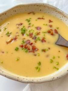 A bowl of creamy yellow vegetable soup topped with crispy bacon bits and chopped chives, with a dark spoon resting in the creamy vegetable soup.