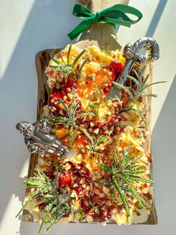 A festive Holiday Butter Board topped with nuts, fruit preserves, herbs, and sugared rosemary sprigs, arranged on a wooden board with two decorative cheese knives and a green ribbon bow.