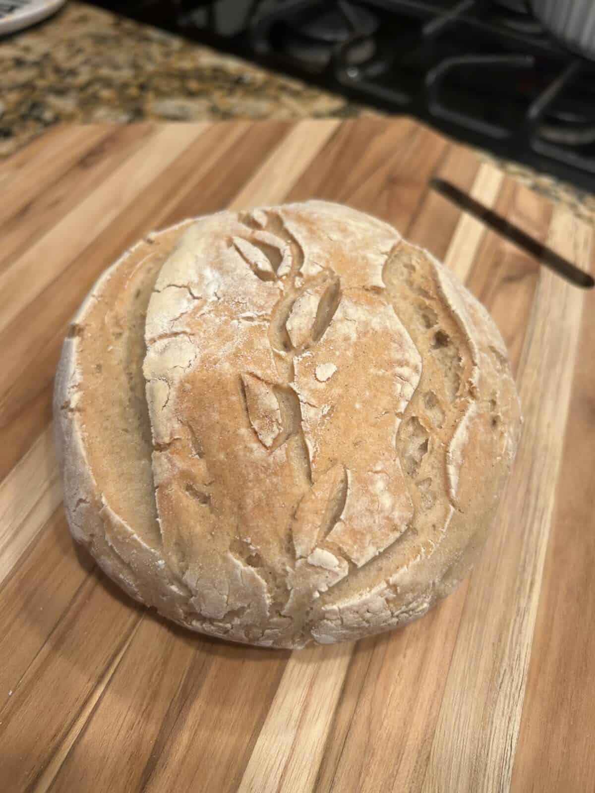 A round loaf of rustic artisan bread with a golden crust and decorative scoring rests on a wooden cutting board in a kitchen setting.