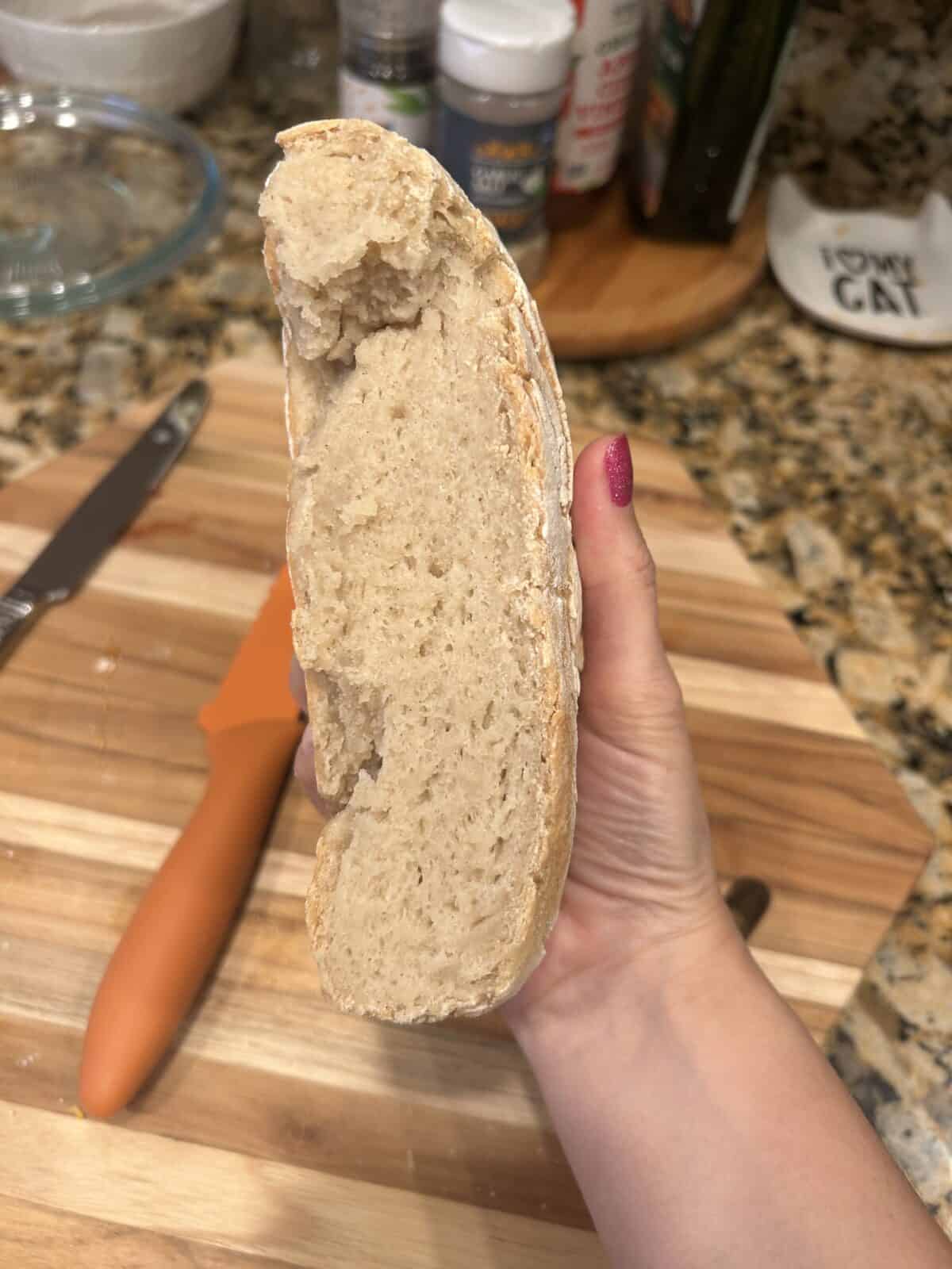 A hand with pink-painted nails holds a sliced piece of rustic bread, revealing its soft and airy crumb. The background shows a wooden cutting board, a knife, a spatula, and kitchen items.