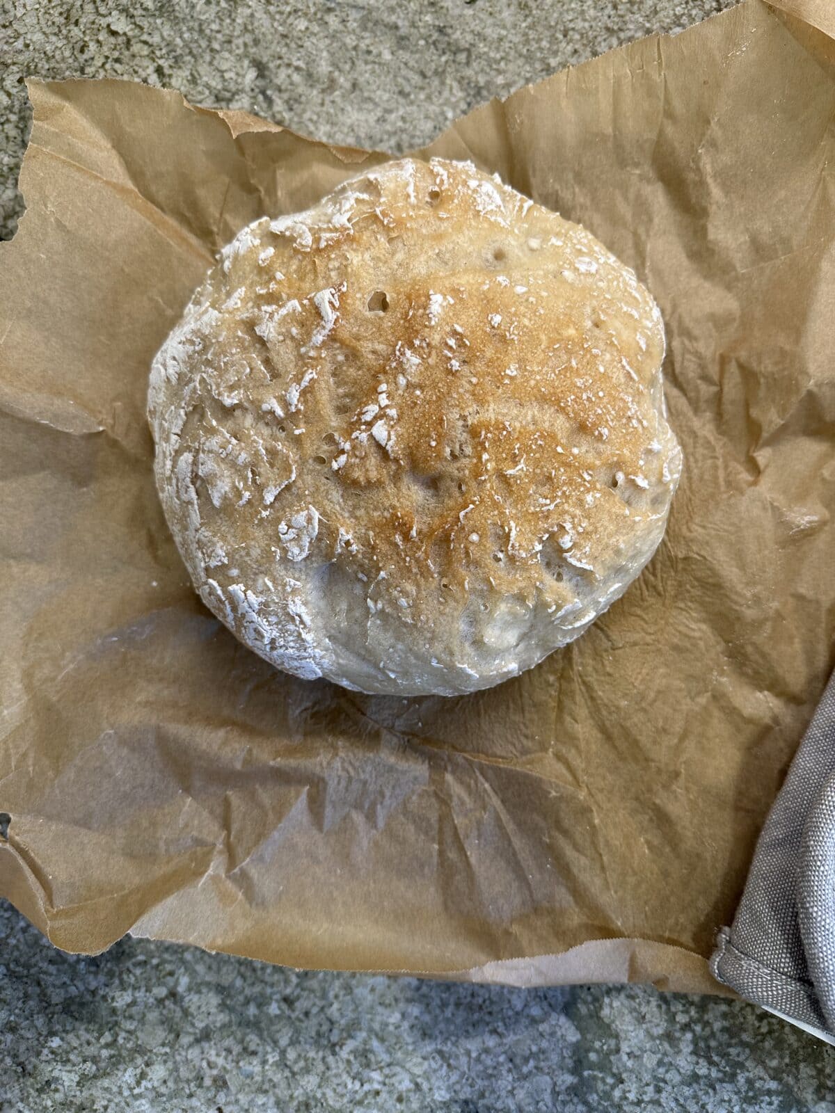 A round loaf of rustic bread with a lightly floured crust sits on crumpled brown parchment paper on a gray stone surface.