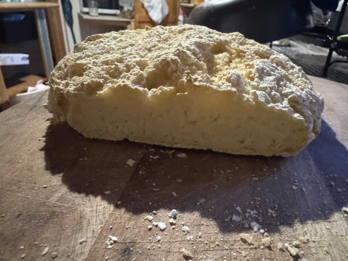 A loaf of no knead gluten free bread with a rough, golden crust and a soft, airy interior sits on a wooden surface in a cozy kitchen setting.