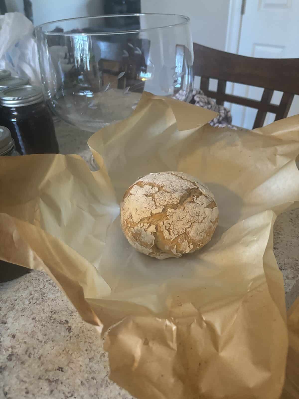 A round loaf of rustic bread sits on crinkled brown paper on a kitchen countertop. A large glass bowl is visible in the background, alongside some jars. The setting suggests a cozy, homey atmosphere.