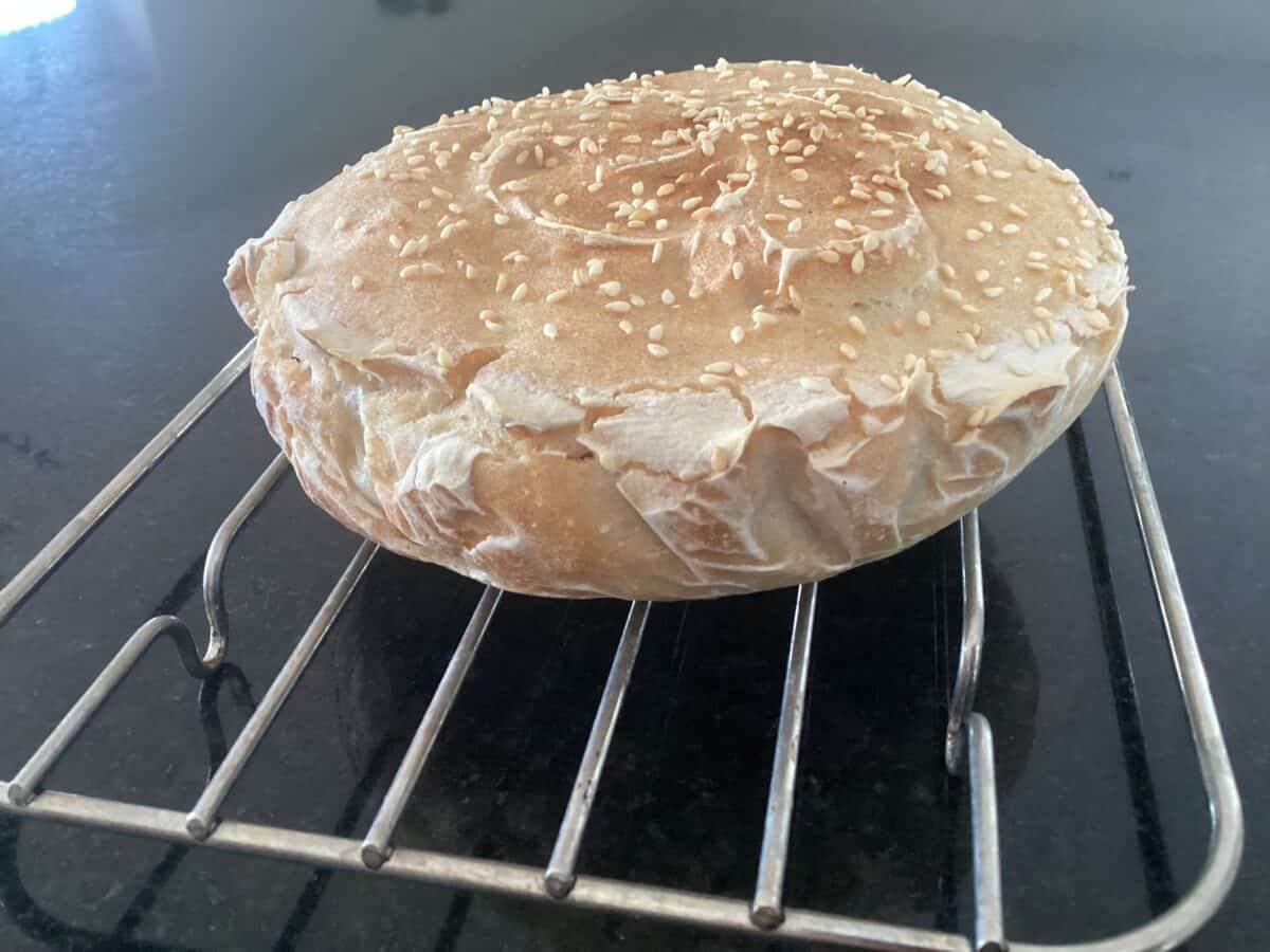 A round, golden-brown loaf of No Knead Gluten Free Bread topped with sesame seeds rests on a metal cooling rack over a dark countertop.