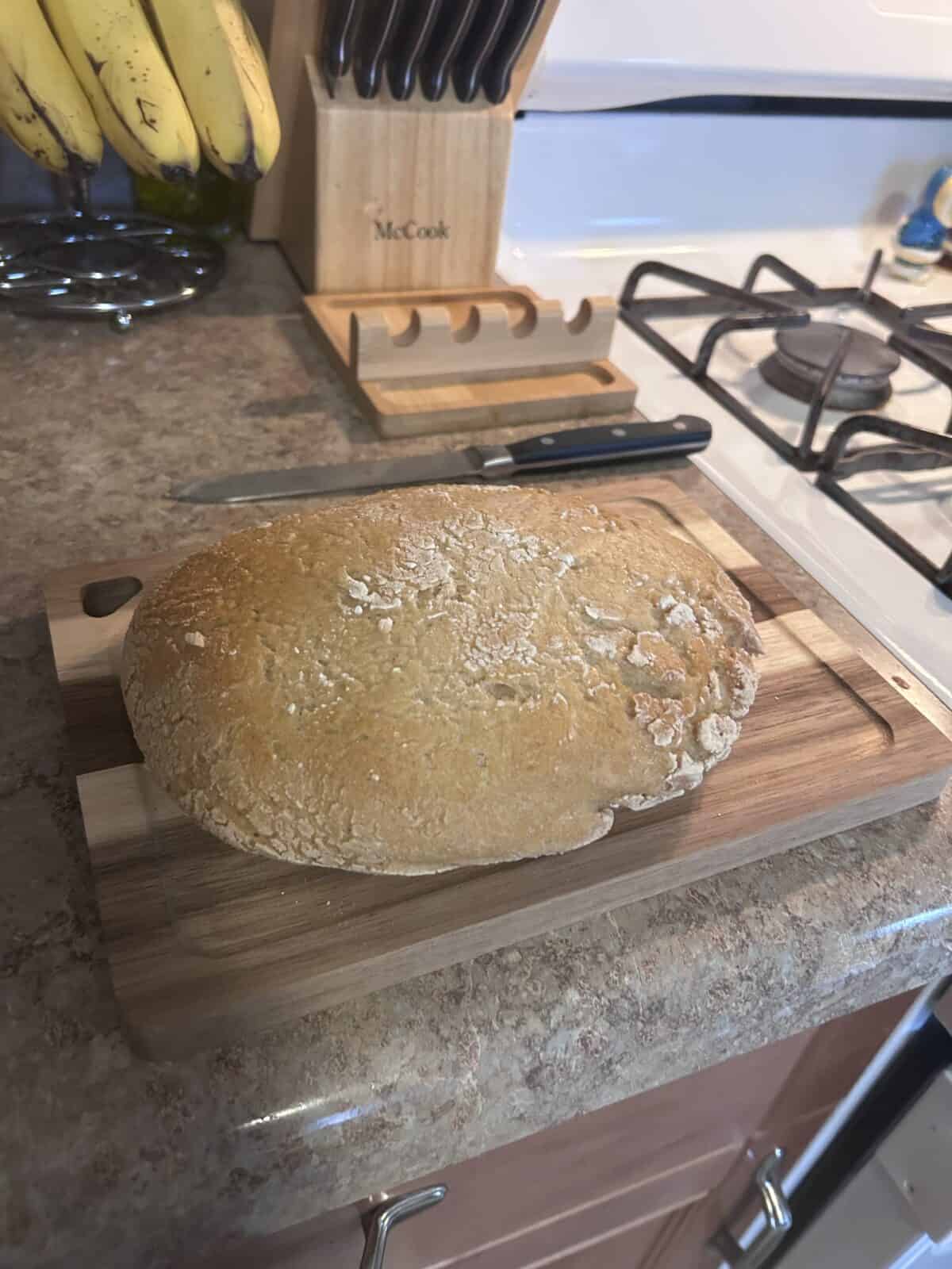A round loaf of No Knead Gluten Free Bread sits on a wooden cutting board on a kitchen counter. A knife rests nearby, with bananas, knives in a block, and part of a gas stove visible in the background.