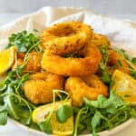 A plate of golden-brown breaded onion rings on a bed of fresh arugula, garnished with lemon wedges.