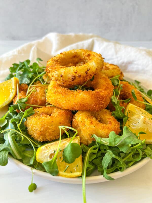A plate of golden-brown breaded onion rings on a bed of fresh arugula, garnished with lemon wedges.