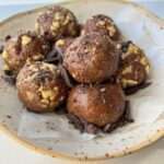 A plate with seven chocolate Energy Balls rolled in chopped nuts, resting on parchment paper and sprinkled with chocolate shavings.
