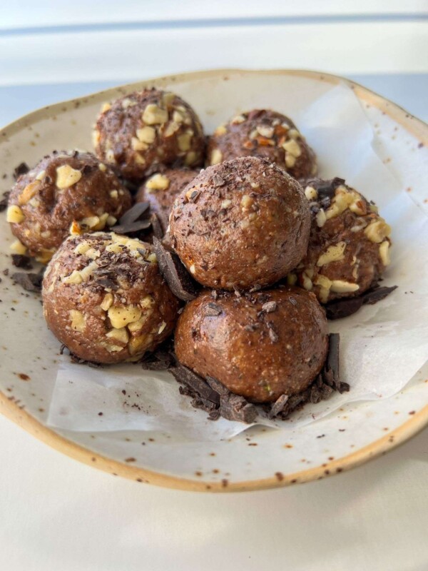 A plate with seven chocolate Energy Balls rolled in chopped nuts, resting on parchment paper and sprinkled with chocolate shavings.