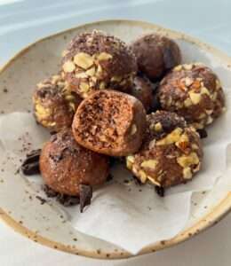 A plate with several chocolate Energy Balls topped with chopped nuts, one bite revealing a dense, fudgy interior. Pieces of chocolate are scattered around the bites on white parchment paper.