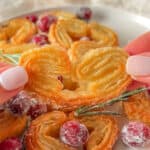 A close-up of a hand holding a heart-shaped Puff Pastry Cookie, with more cookies, sugared cranberries, and rosemary arranged on a plate in the background.
