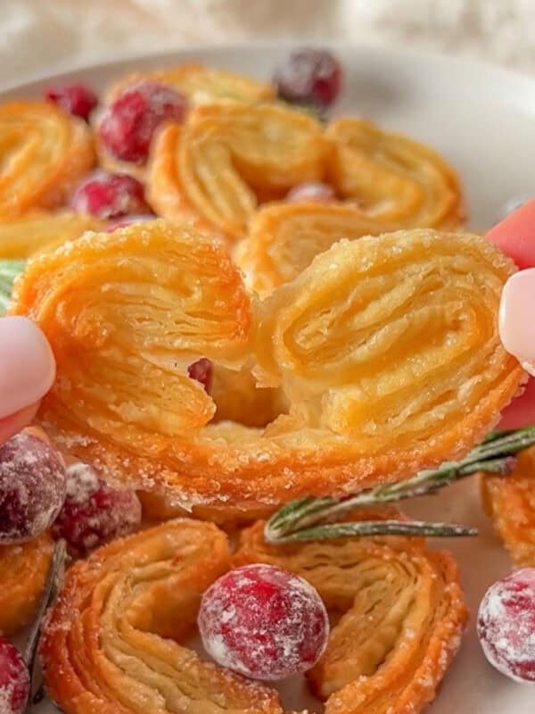 A close-up of a hand holding a heart-shaped Puff Pastry Cookie, with more cookies, sugared cranberries, and rosemary arranged on a plate in the background.