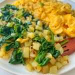 A plate with scrambled eggs and a side of Potato Hash and Spinach Breakfast. The food is brightly colored and looks freshly cooked, featuring sautéed spinach and diced potatoes for a wholesome start to your day.