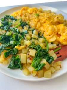 A plate with scrambled eggs and a side of Potato Hash and Spinach Breakfast. The food is brightly colored and looks freshly cooked, featuring sautéed spinach and diced potatoes for a wholesome start to your day.