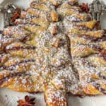 A Puff Pastry Christmas Tree dusted with powdered sugar sits on a white plate, garnished with star anise and festive silver napkin rings on each side.