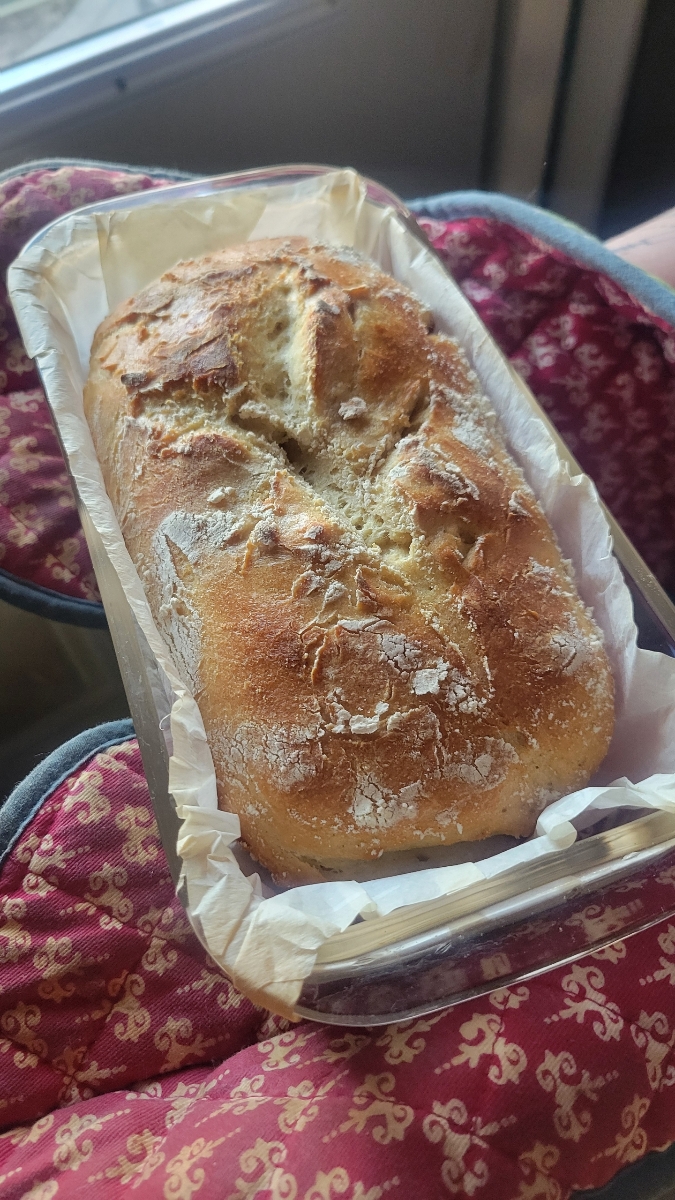 A freshly baked loaf of No Knead Gluten Free Bread rests in a parchment-lined loaf pan, held by someone wearing red oven mitts with a yellow pattern.