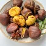 A ceramic bowl filled with roasted chestnuts, some peeled to reveal their yellow insides, is garnished with a sprig of rosemary and a star anise, all set against a white background.