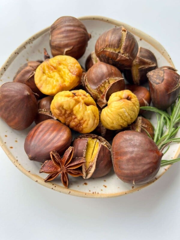 A ceramic bowl filled with roasted chestnuts, some peeled to reveal their yellow insides, is garnished with a sprig of rosemary and a star anise, all set against a white background.