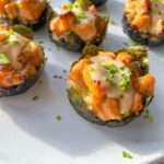 Close-up of several Salmon Sushi Cups on a white plate. Each cup features a nori seaweed base filled with a creamy, diced topping and is garnished with chopped chives. Sunlight shines on the plate.