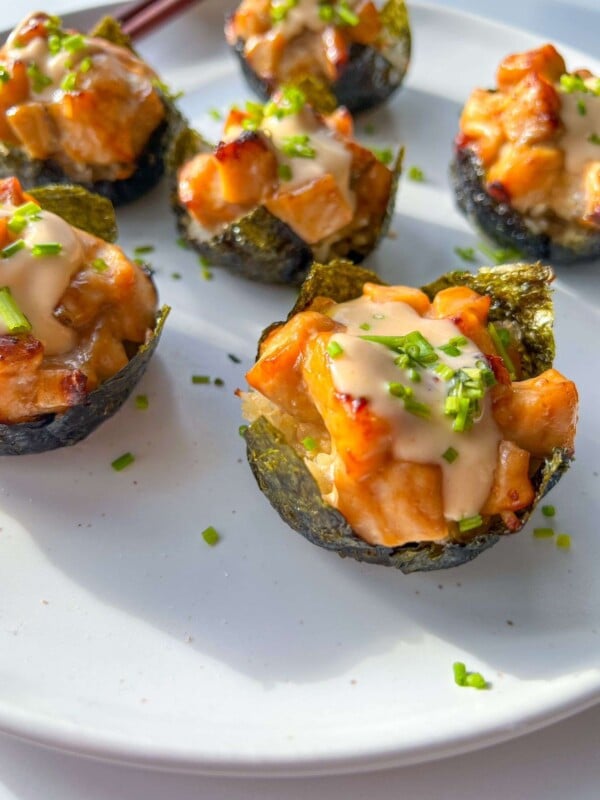 Close-up of several Salmon Sushi Cups on a white plate. Each cup features a nori seaweed base filled with a creamy, diced topping and is garnished with chopped chives. Sunlight shines on the plate.