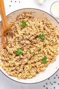 A large white dish filled with rotini pasta tossed in a creamy sauce, garnished with fresh parsley. A wooden spoon rests on the side. Black peppercorns are scattered on the table, and a small bowl of grated cheese is nearby.