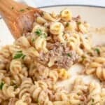 A wooden spoon lifts a serving of creamy rotini pasta mixed with shredded meat, garnished with chopped parsley. The pasta is in a white dish, and a sprig of parsley is visible to the side.