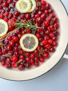 A white pot filled with fresh cranberries, lemon slices, star anise, a cinnamon stick, and a sprig of rosemary in water—perfect for creating homemade Holiday Potpourri.
