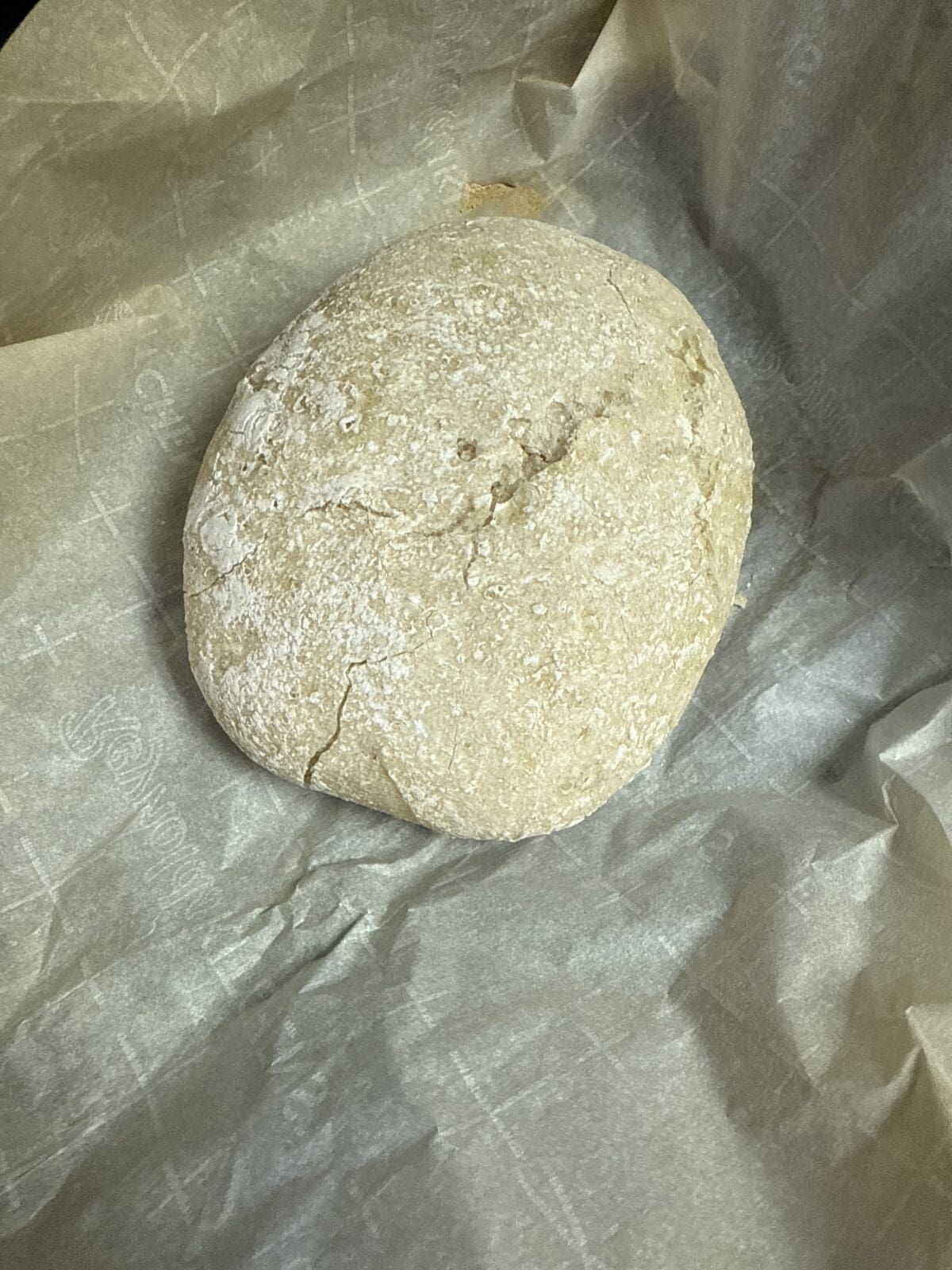 A round loaf of rustic, lightly floured No Knead Gluten Free Bread sits on a sheet of parchment paper with a faint grid pattern. The bread has a slightly cracked, golden-brown crust.