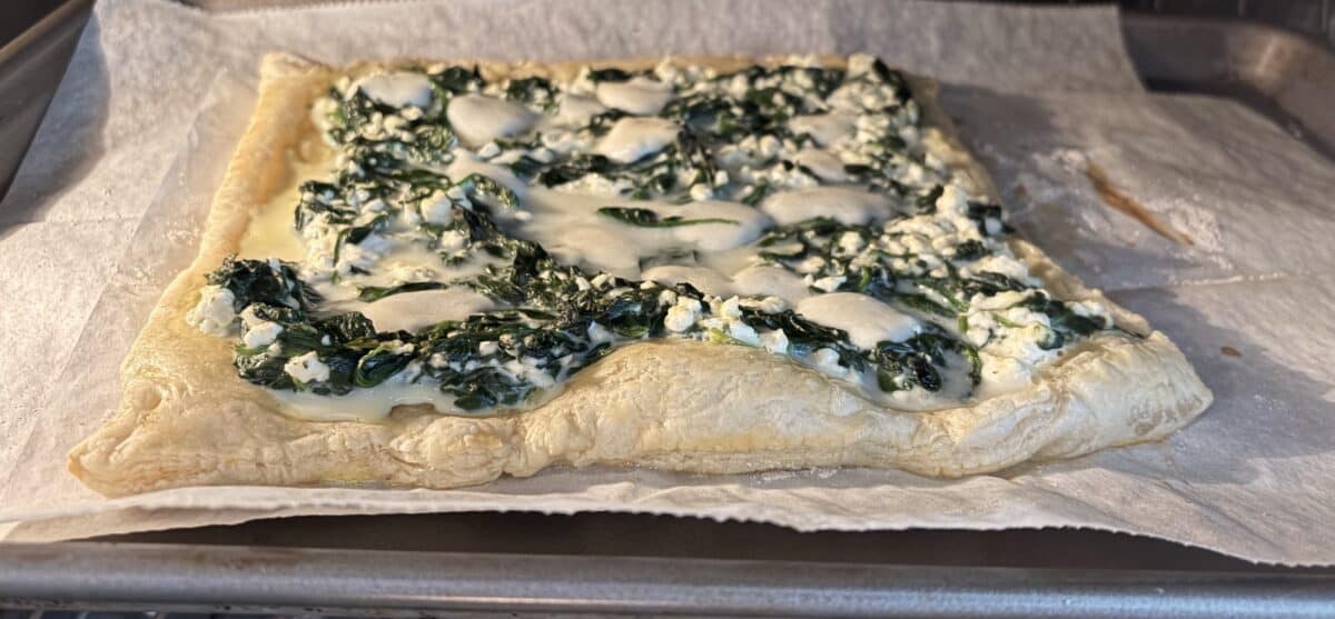A square Puff Pastry Spanakopita tart is baking on parchment paper in a metal tray, with melted cheese and spinach filling spread evenly over the golden, flaky crust.