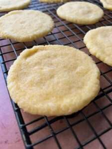 Close-up of 3 Ingredient Sugar Cookies cooling on a black wire rack. The cookies are round, golden, and have a slightly rough texture from sugar crystals. A pinkish surface is visible in the background.
