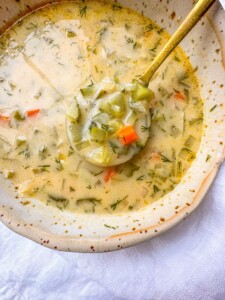 A close-up of creamy dill pickle soup with chopped carrots, celery, potatoes, and fresh herbs, served in a rustic bowl with a golden ladle lifting a spoonful.