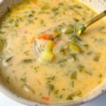 A creamy dill pickle soup with chopped carrots, potatoes, pickles, and herbs served in a speckled ceramic bowl with a golden spoon.